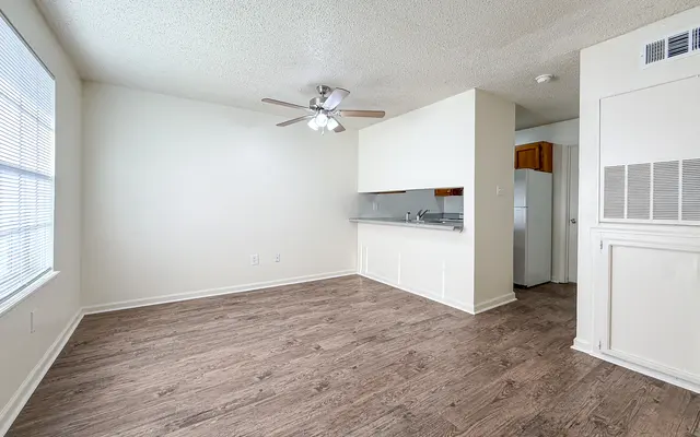 A spacious and empty apartment living room with light-colored walls and wooden flooring. It features a ceiling fan and a kitchen area in the background, which includes a refrigerator and a countertop. Large windows allow natural light to fill the room.
