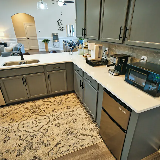 A modern kitchen featuring gray cabinetry, a white countertop, and various appliances including a coffee maker and microwave. The kitchen is complemented by a decorative rug and has a spacious layout visible in the background.