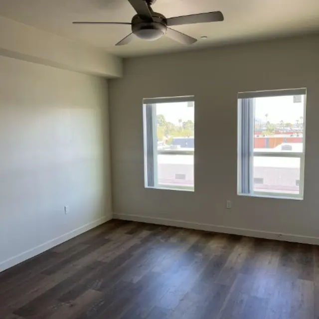 A spacious empty room with two windows and a ceiling fan, featuring hardwood flooring and light-colored walls.
