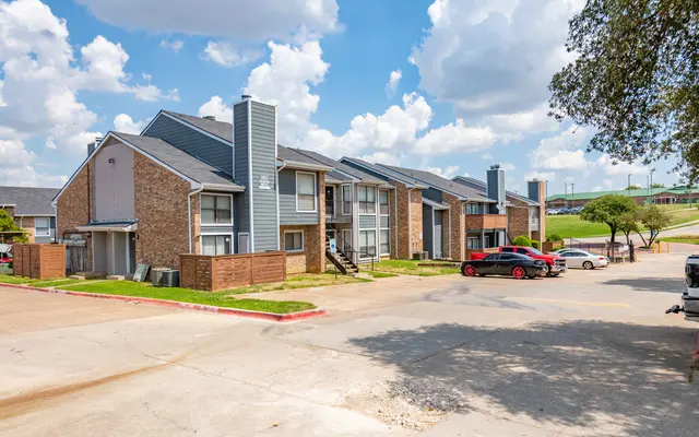 View of an apartment complex with two-story buildings, featuring a mix of brick and siding exteriors, set against a partly cloudy sky.