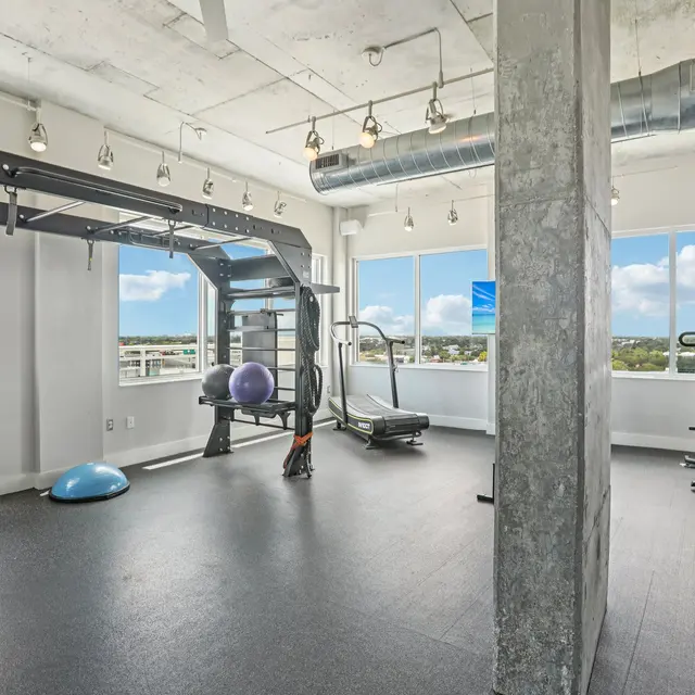 A modern gym interior featuring various workout equipment including a punching bag, dumbbells, exercise balls, and cardio machines in a bright, airy setting with large windows and a view of the outdoors.