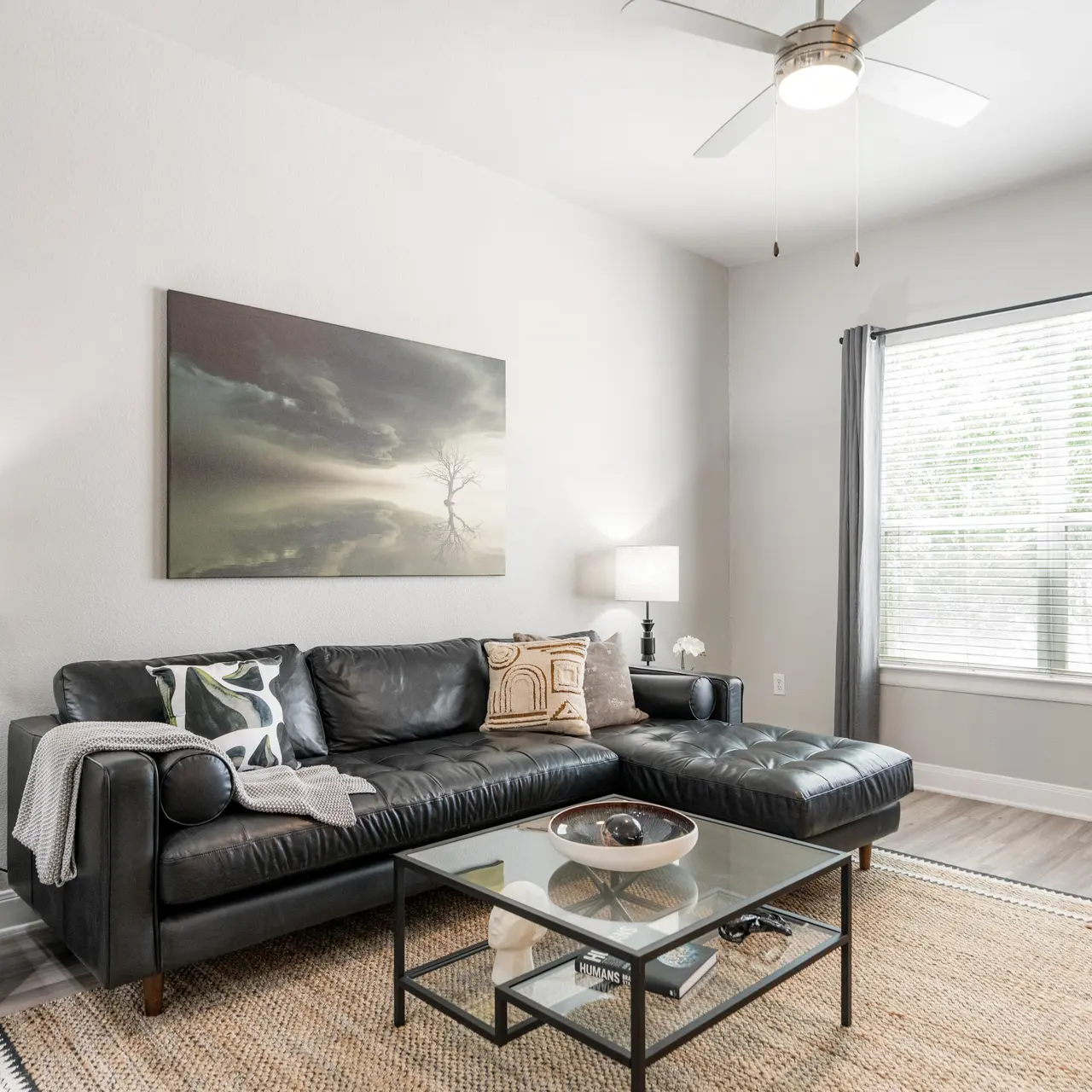 A contemporary living room featuring a black leather sectional sofa, a glass coffee table, and two lamps. The walls are painted in soft colors, and there is a large painting on the wall. Natural light streams in through a window adorned with curtains, and a woven rug covers the floor.