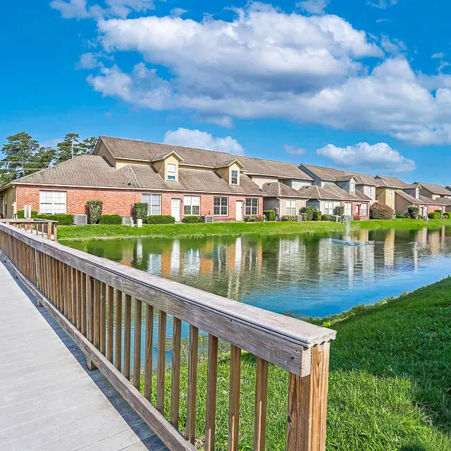 A scenic view of an apartment complex along a calm water body, with a wooden walkway leading towards the buildings.