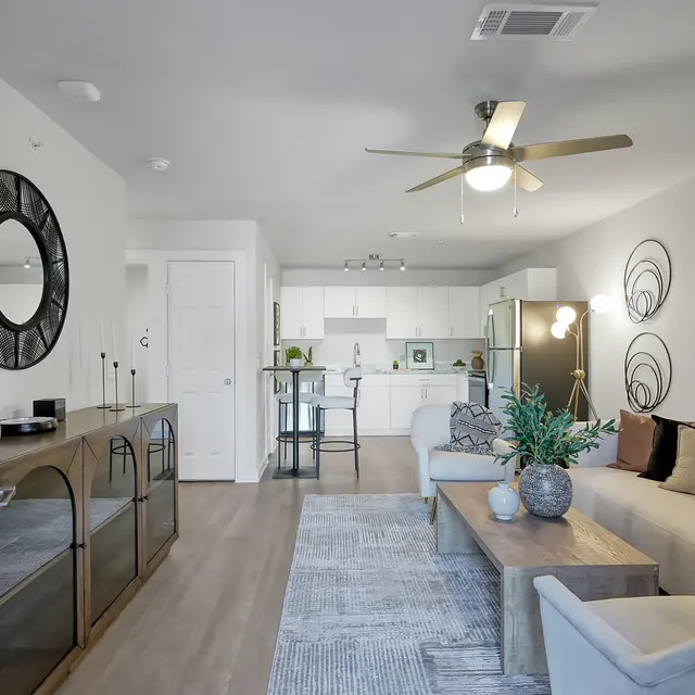 A modern living room with a beige sofa, circular mirror, and a coffee table with plants. The room has a well-lit kitchen in the background, with a dining table, and light wooden flooring.