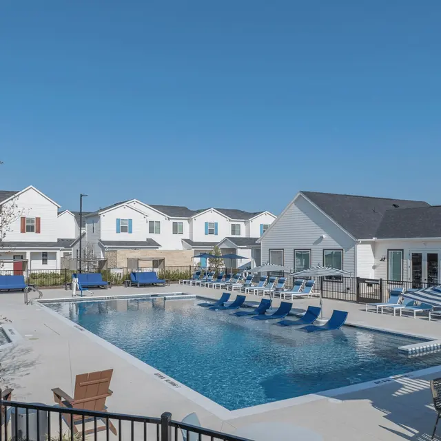 A sunny day at a residential apartment complex featuring a large swimming pool with lounge chairs around it and white buildings in the background.