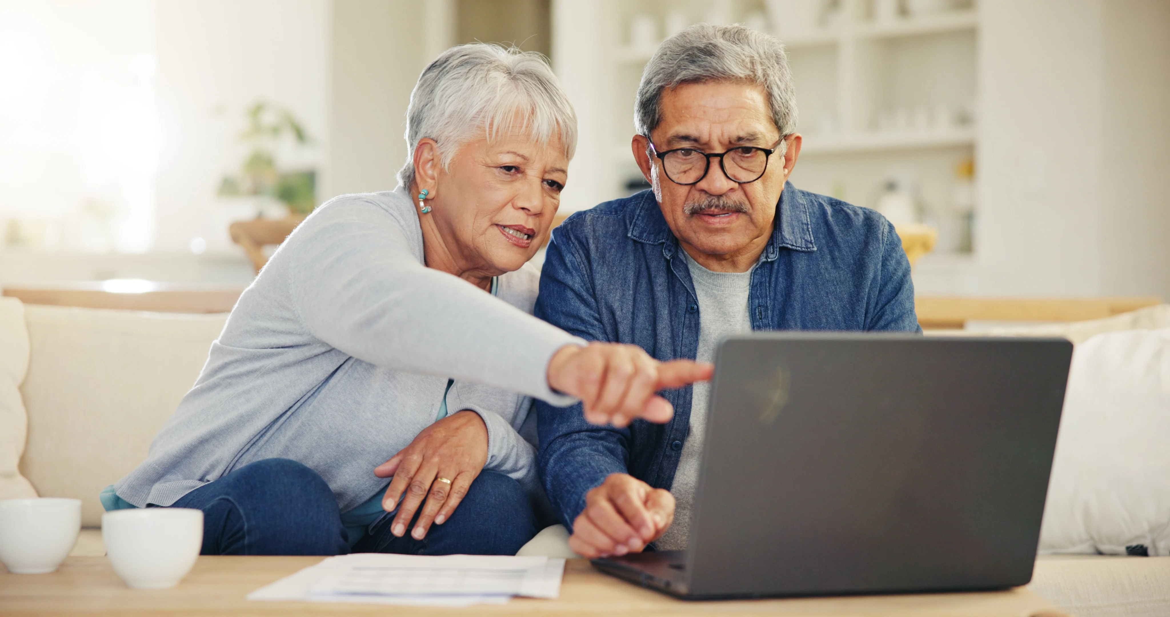 An elderly couple sitting on a couch, looking intently at a laptop. One is pointing at the screen while the other is observing. They appear engaged and focused.