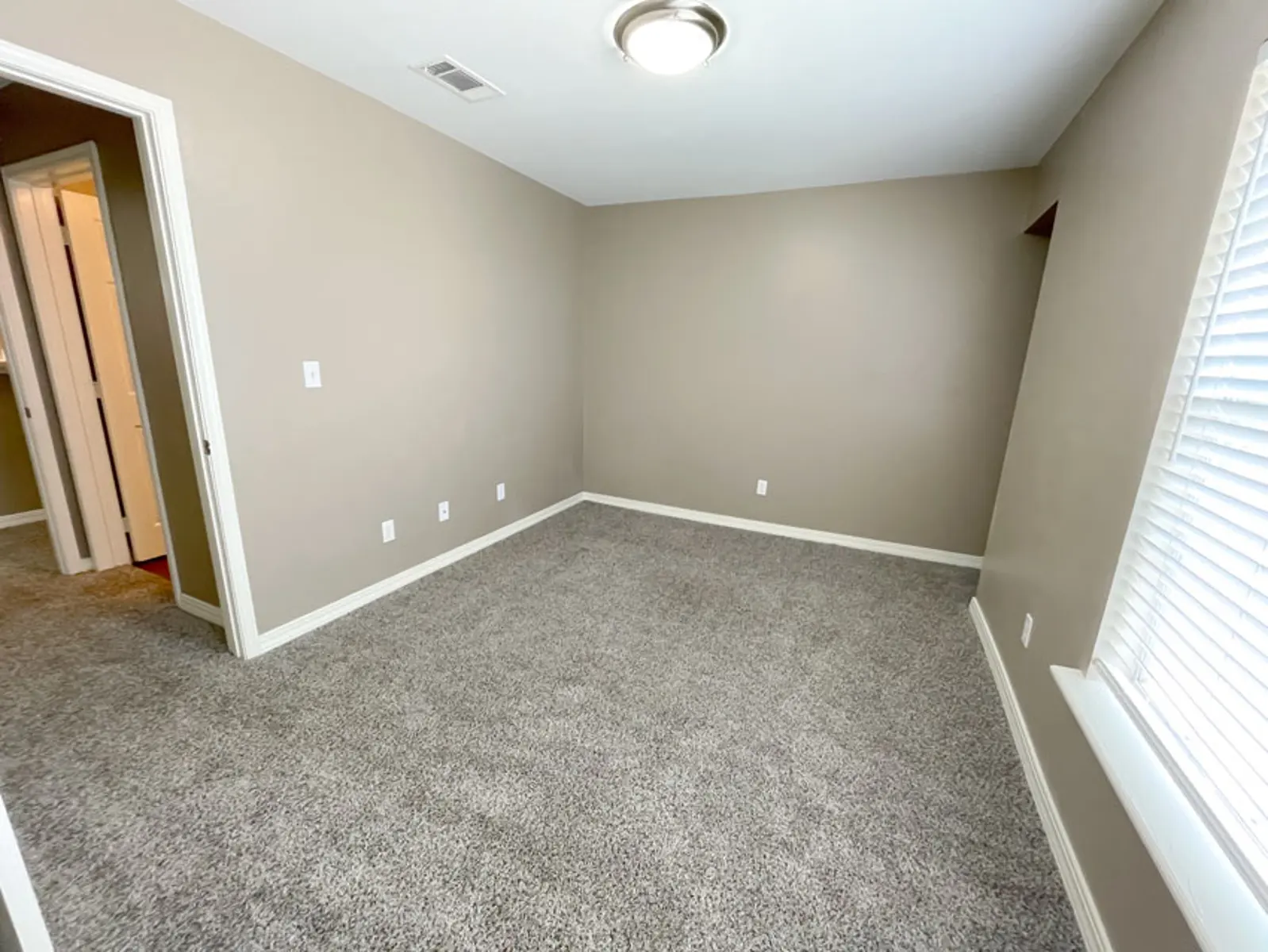 Empty bedroom with beige walls and carpet, featuring a window and a door leading to another room.