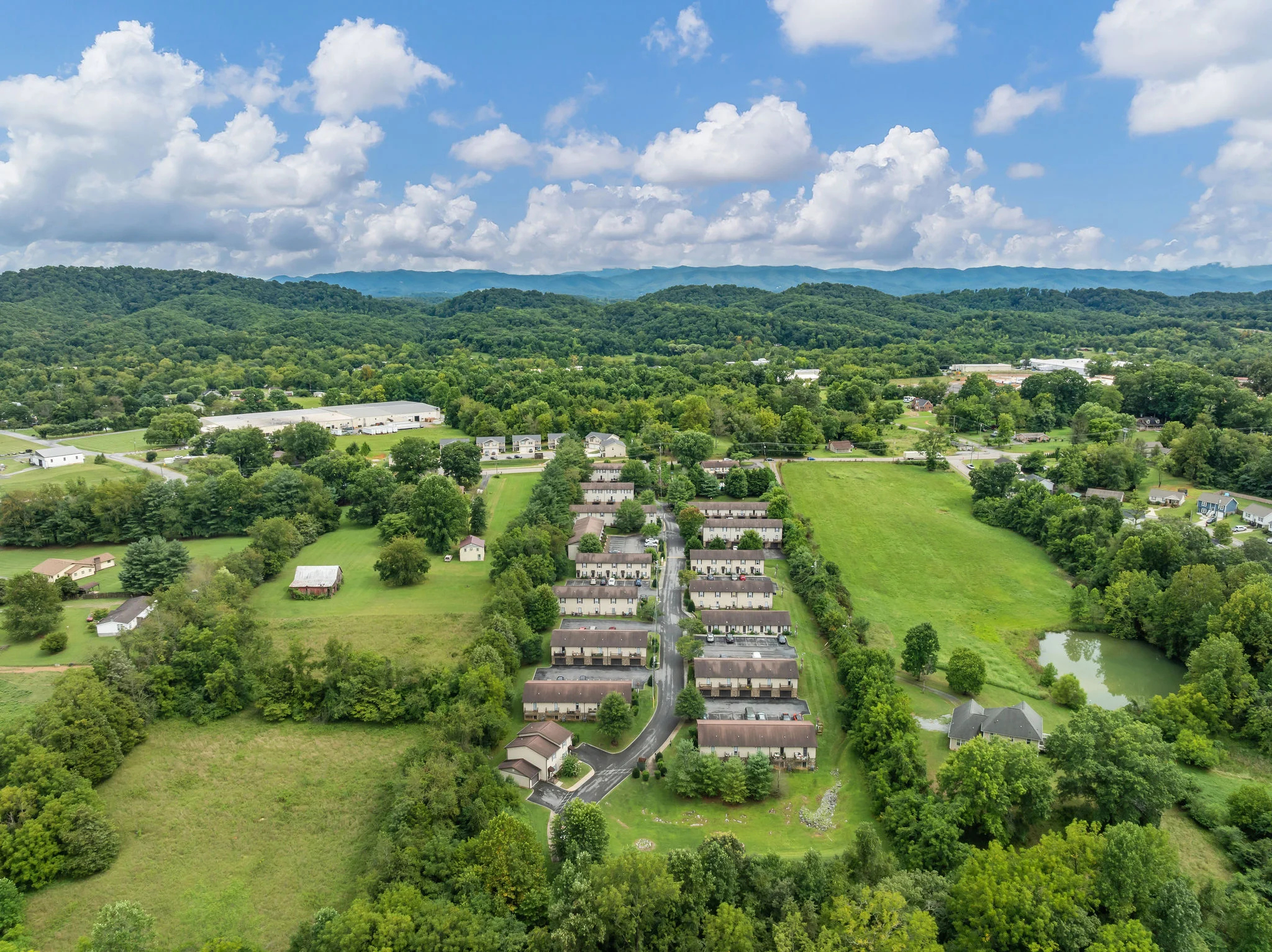 Aerial view of a residential complex surrounded by green landscapes and hills under a partly cloudy sky.