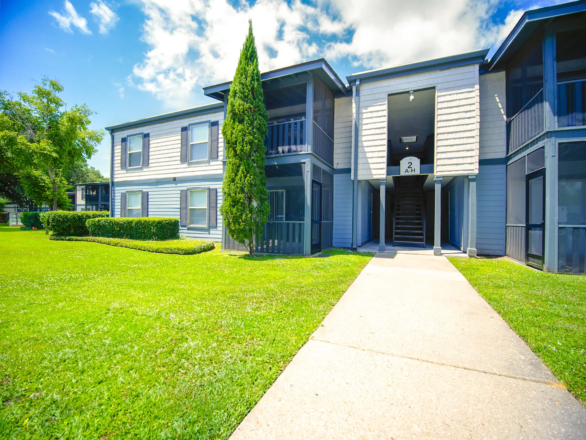 Exterior view of a two-story apartment building with green lawn and trees under a blue sky.