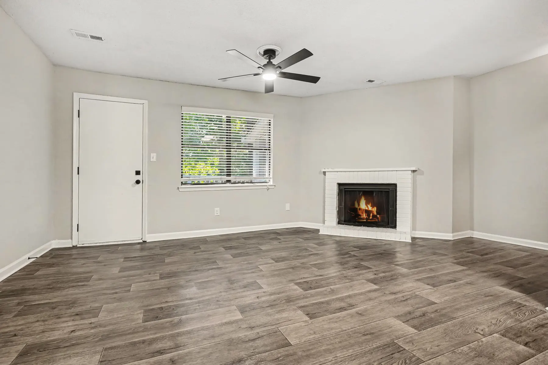 A spacious living room featuring a fireplace, hardwood-style flooring, and a ceiling fan. The walls are painted in a neutral shade, and there is a window with blinds letting in natural light.