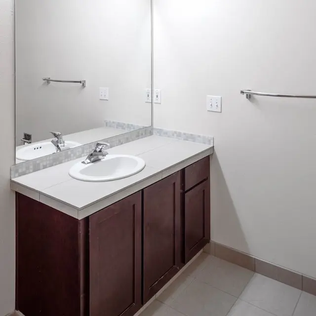 A clean, modern bathroom with a white sink, faucet, and a large mirror. There is a low wooden cabinet underneath the sink and a towel rack on the wall.