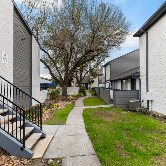 Apartment Complex Walkway A walkway in an apartment complex lined with greenery and trees, featuring buildings with stairs and a light gray facade.