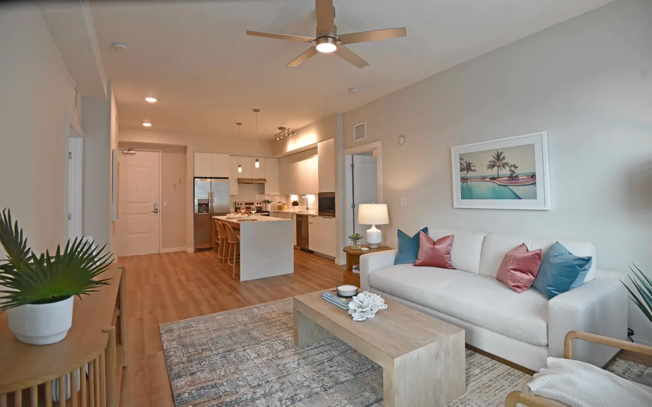 Modern living room with a cream-colored sofa, decorative pillows, a wooden coffee table, and plants. The space is open to a kitchen area with bar seating.