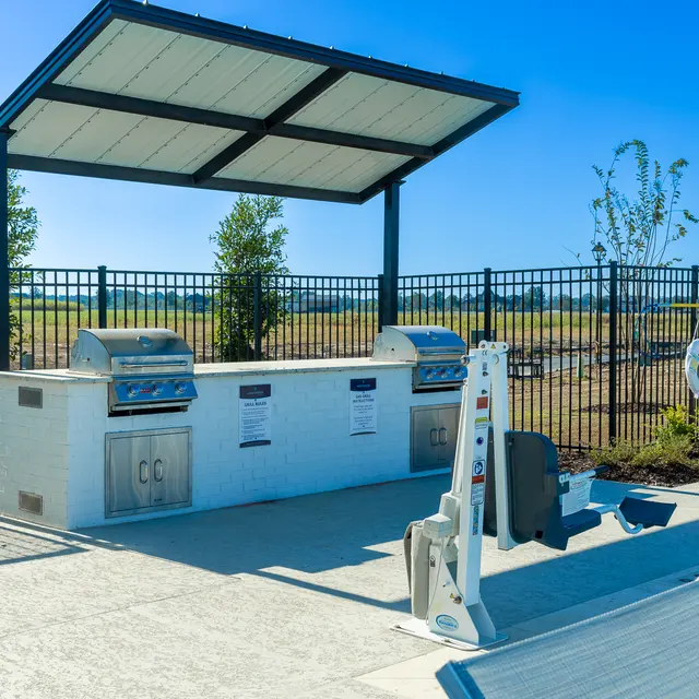 Outdoor grilling area next to a swimming pool with two grills and shaded seating.
