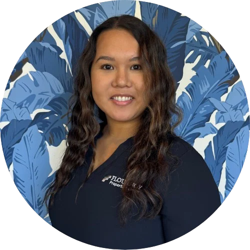 A smiling woman with long wavy hair, wearing a dark collared shirt, stands in front of a blue patterned background featuring large leaves.