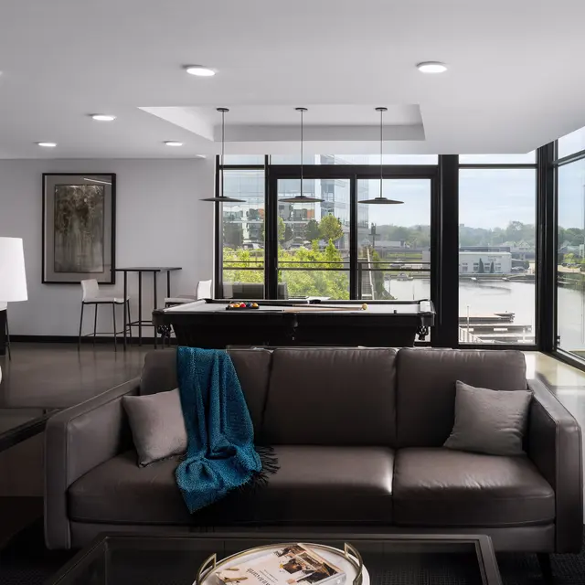 A modern living room featuring large windows with a view of a river, a brown leather sofa, stylish lamps, and minimalist decor in a well-lit space.