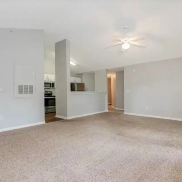 A spacious living room featuring beige carpeting, a ceiling fan, and an open layout connecting to a kitchen area. The walls are painted in a light gray color, and there is a doorway leading to a bathroom visible in the background.