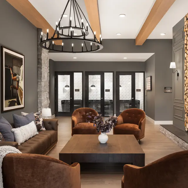 A stylish modern living room featuring comfortable brown chairs and a brown sofa, a coffee table at the center, and framed artwork on the walls. A stone fireplace is on the right side, and large windows with black frames are at the back, allowing natural light to enter.