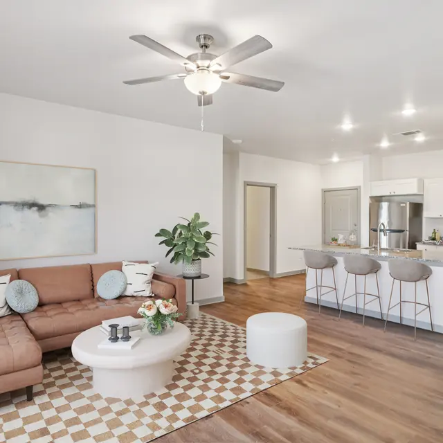 A modern living room with a brown leather sectional sofa, round coffee table, and decorative rug. The kitchen is visible in the background with white cabinetry and bar stools.