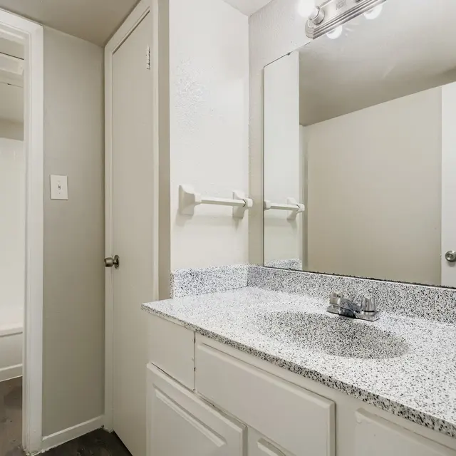 Interior view of a bathroom featuring a granite countertop with a sink, a large mirror, and a door leading to a shower area.