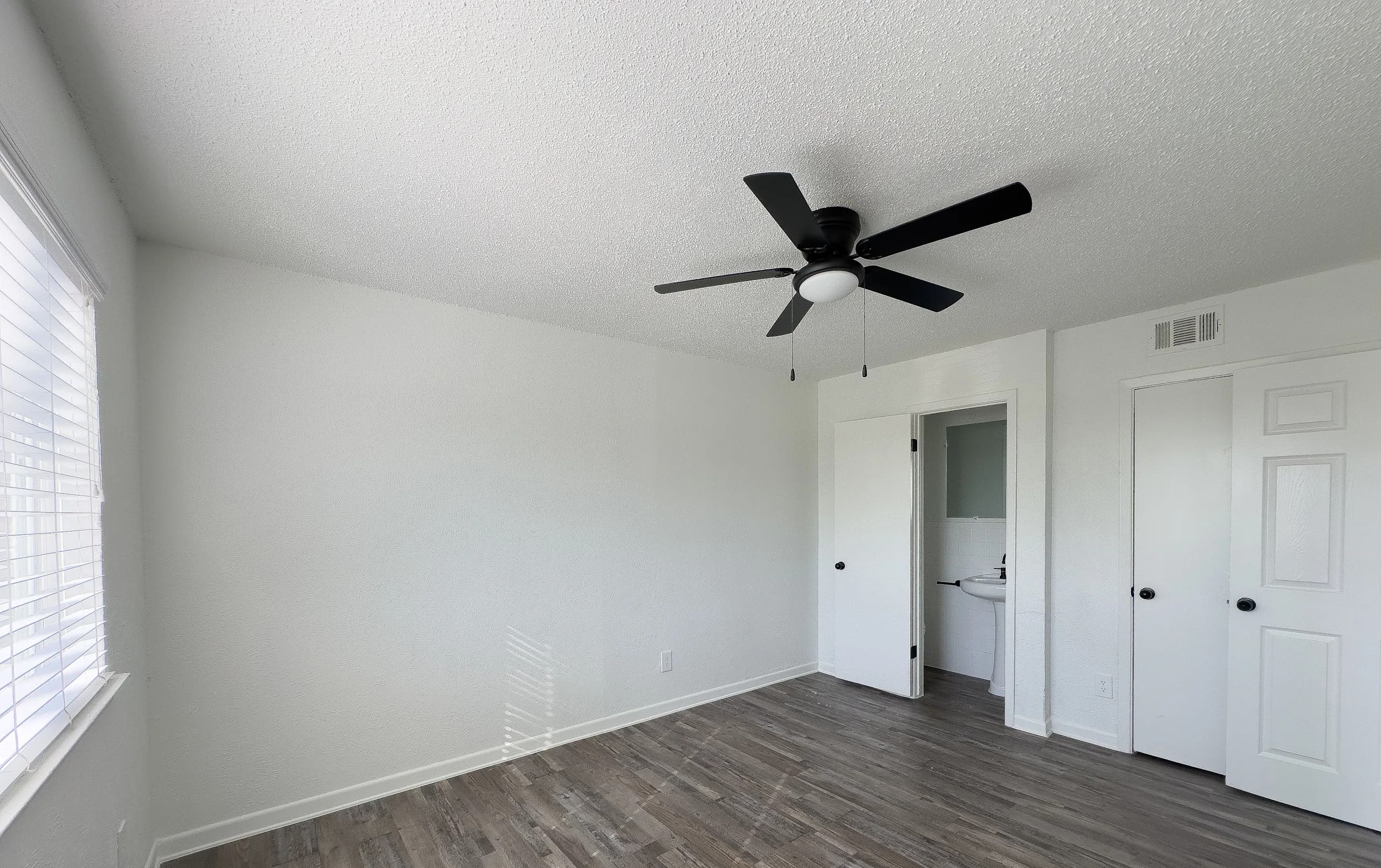 Modern Bedroom Interior Interior view of a modern bedroom with light-colored walls, wooden flooring, and a ceiling fan. A window allows natural light, and a door leads to a bathroom area.