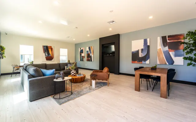 A spacious and modern living room featuring a grey sectional sofa, wooden coffee table, and a dining table. Two large abstract paintings hang on the wall, and a small indoor plant is placed near the sofa. The room has light wooden flooring and a neutral color palette with accent colors.