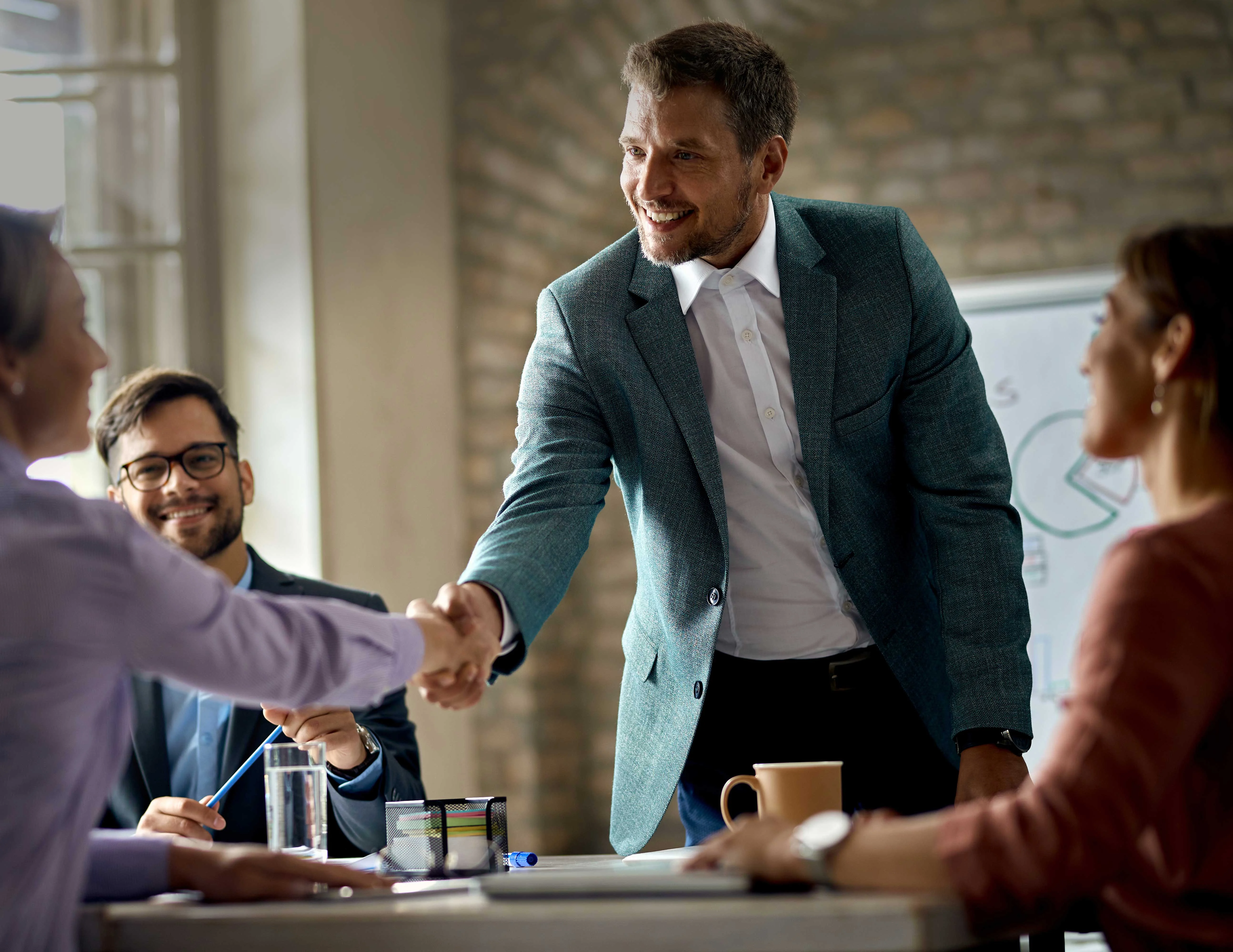 Business Meeting Handshake A man in a teal blazer shakes hands with a woman in a light purple blouse at a business meeting, with another man smiling in the background and a woman seated opposite them.
