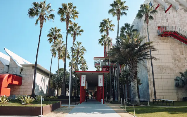 A sunny urban scene featuring a pathway between modern architectural buildings, palm trees lining the walkway, and a large red entrance structure.