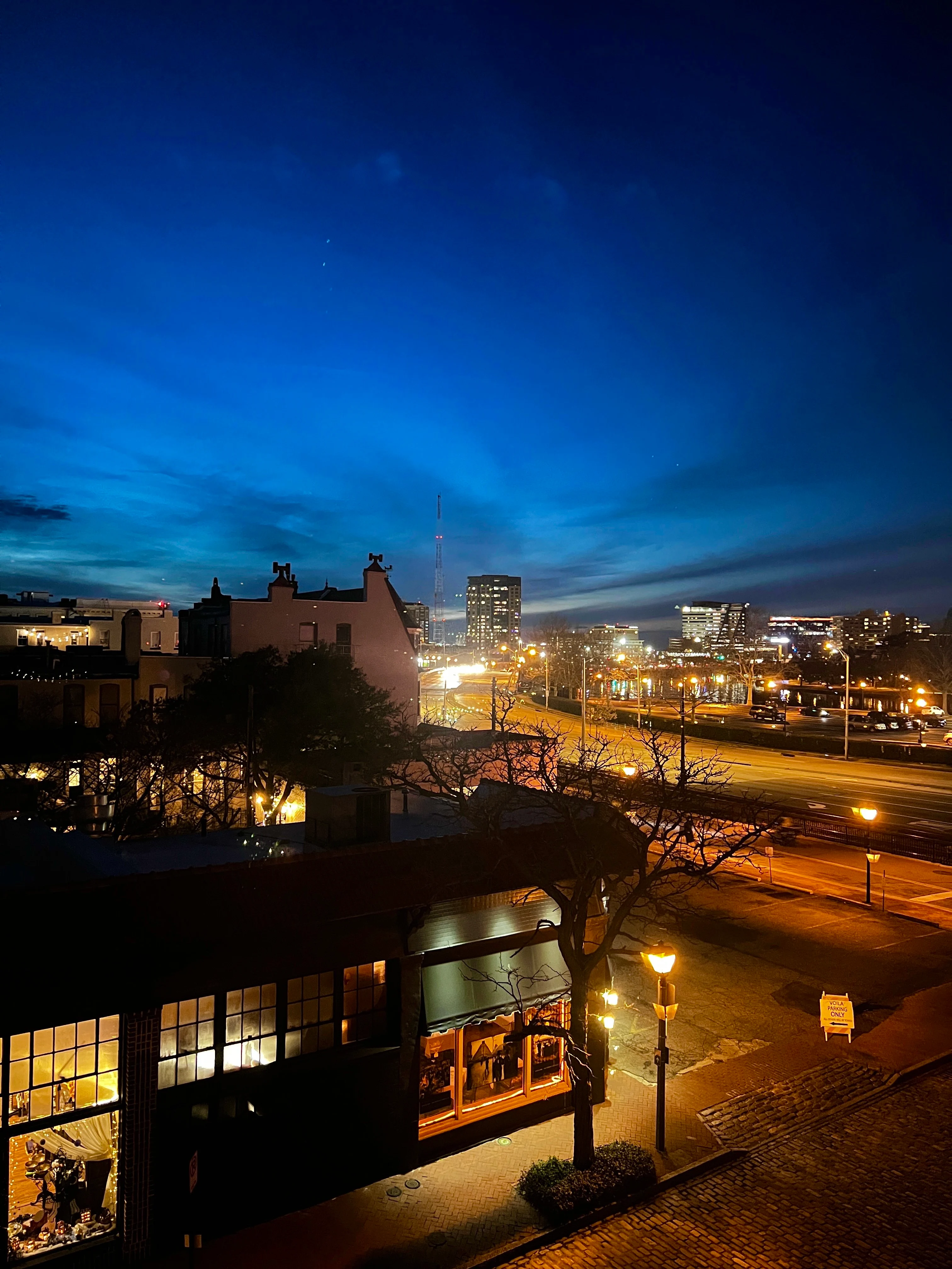 Dusk Cityscape A scenic view of a city at dusk, featuring a darkening blue sky, illuminated buildings, and a calm street with streetlights. A restaurant or shop is visible in the foreground, along with trees and a cobblestone path.