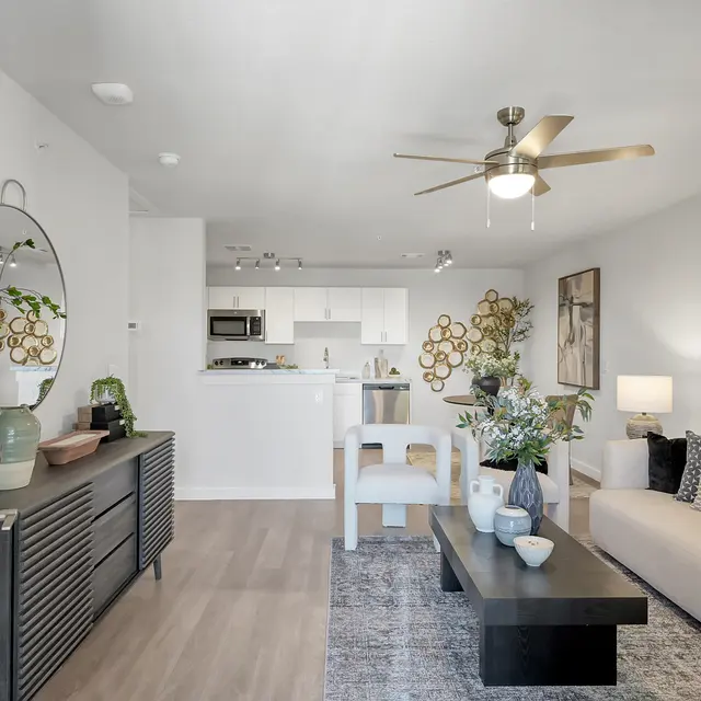 A modern living room featuring a beige sofa, a black coffee table, decorative plants, and artwork on the walls, with a kitchen visible in the background.