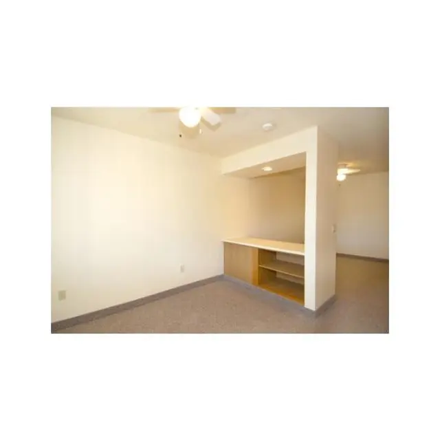 Empty Apartment Room An empty room with beige walls and gray carpeting, featuring a ceiling fan and a half wall with a countertop.