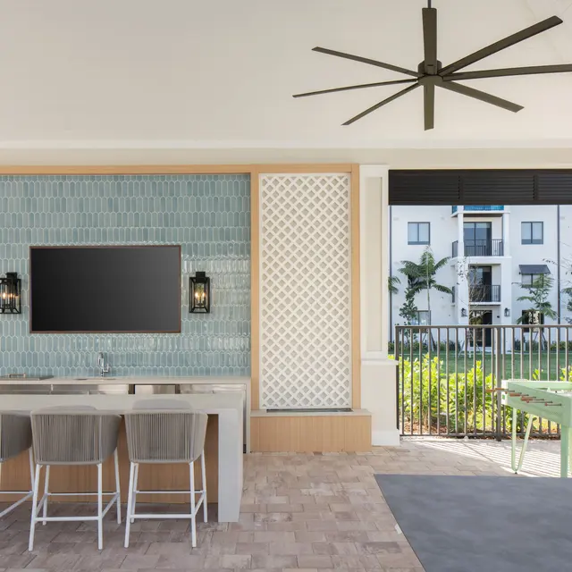 A stylish outdoor lounge area featuring a large flat-screen television mounted on a decorative wall, surrounded by seating at a bar. The space has ceiling fans and a view of greenery through large windows.