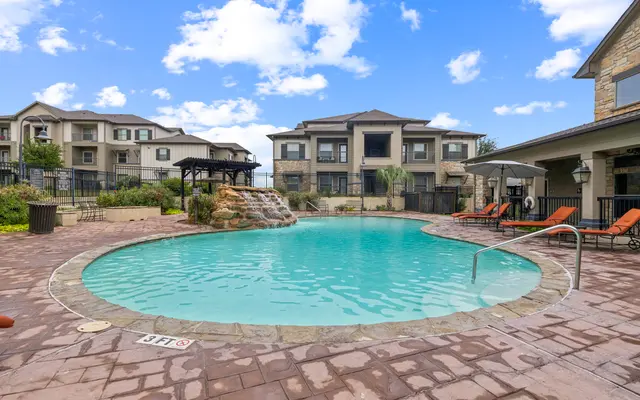 Apartment Pool Area A swimming pool surrounded by a stone patio, featuring a waterfall and lounge chairs, with modern apartment buildings in the background under a blue sky.