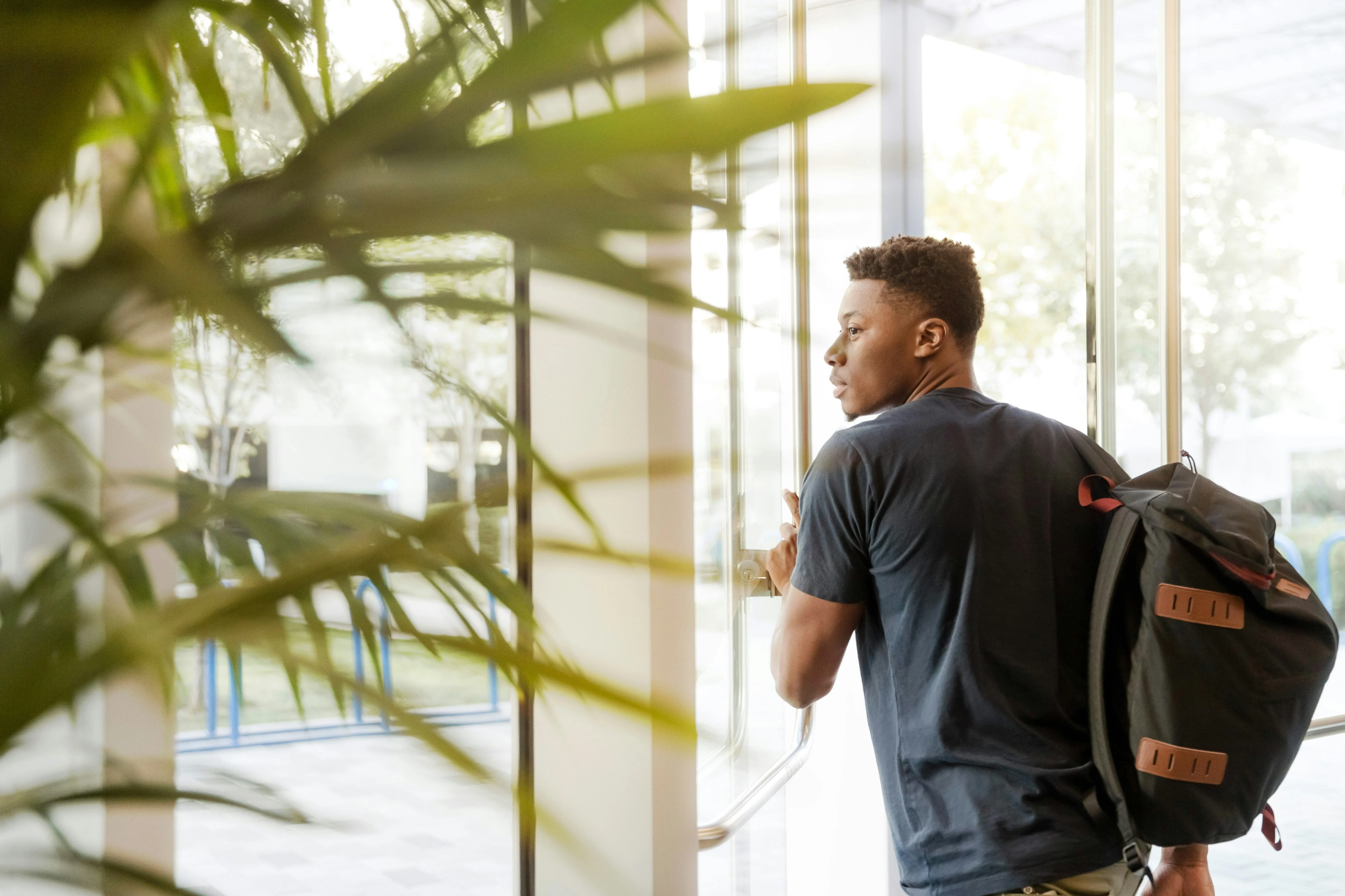 A young man with a backpack is exiting a building, visible through a glass door. He appears focused and is holding the door open with one hand, while looking back. In the foreground, there are green leaves from a plant.