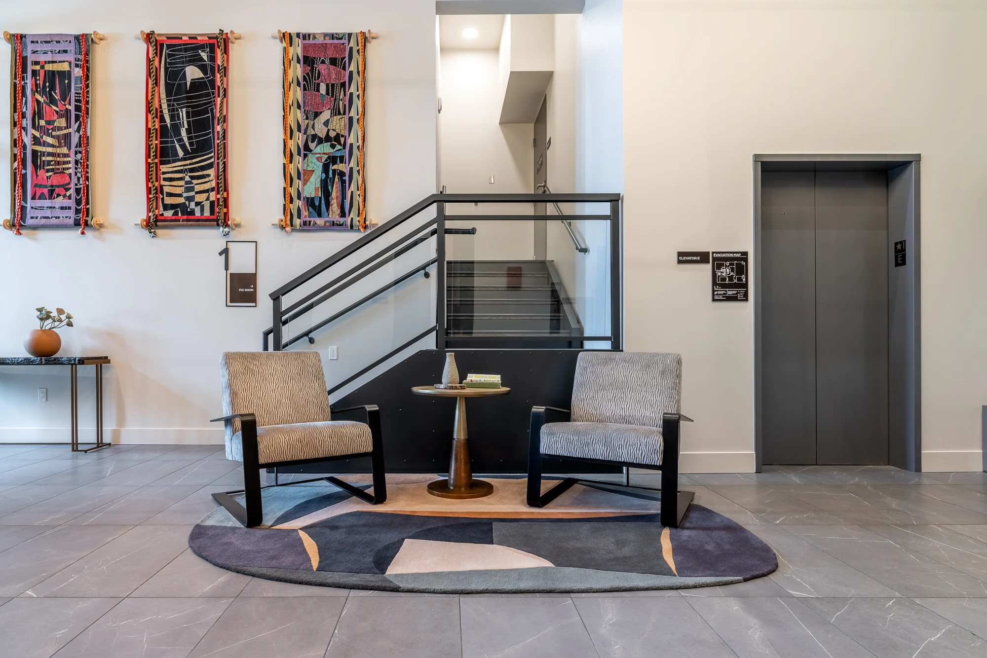 Modern Lobby Design A modern lobby area featuring two gray chairs, a round side table, and artistic wall decor. An elevator door is visible to the right, alongside a staircase with a contemporary railing.