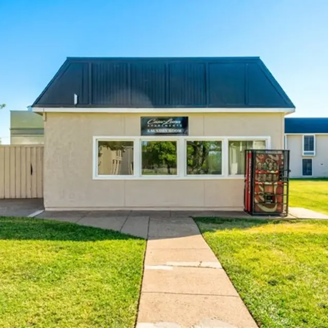 A light-colored building with a black roof and large windows. A vending machine is positioned in front of the building on a paved walkway, surrounded by green grass. There are trees and another building visible in the background under a clear blue sky.