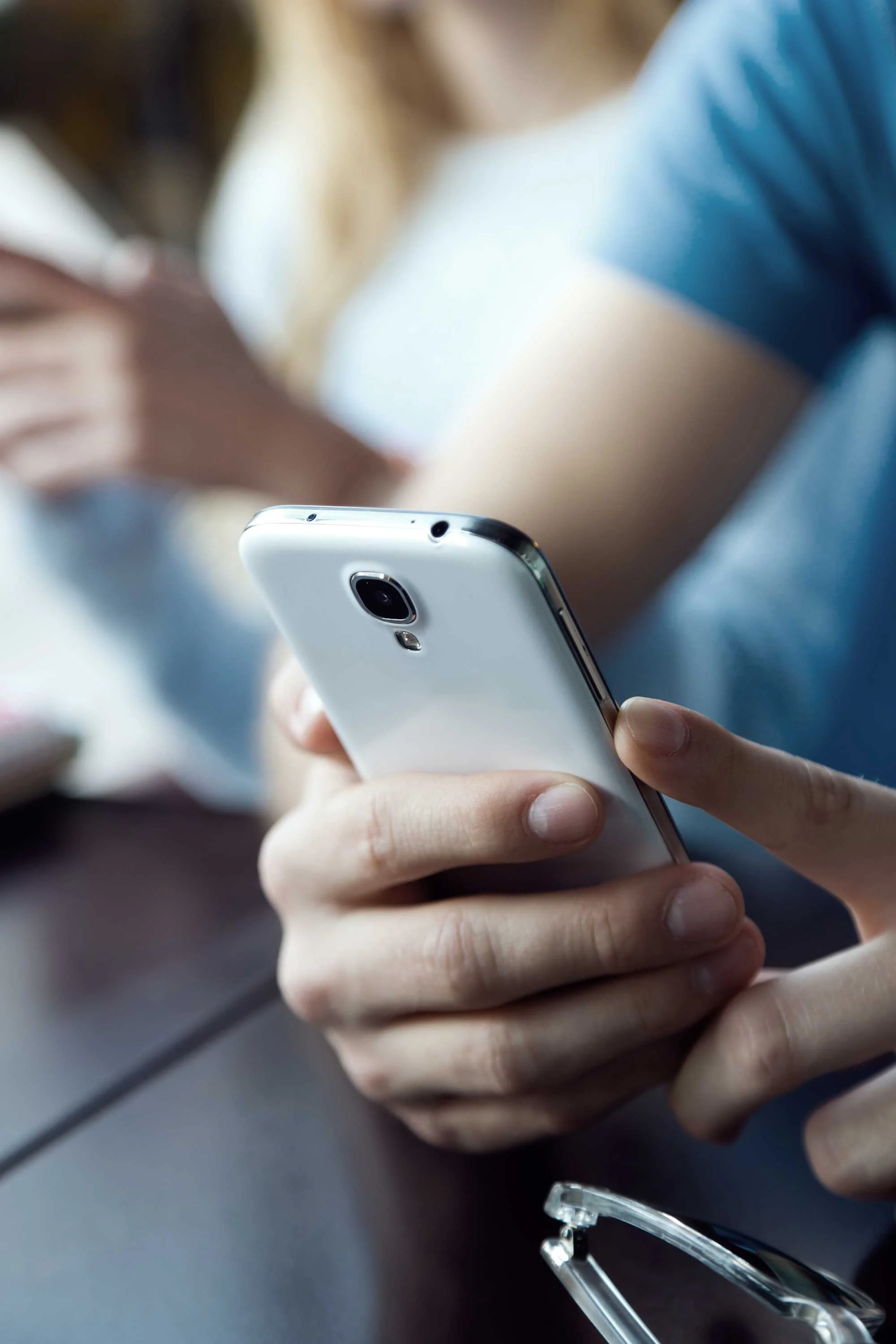Smartphone Use in a Social Setting A close-up of a person's hands holding a smartphone, with another person partially visible in the background, indicating a social setting like a café. The focus is on the smartphone in the foreground.