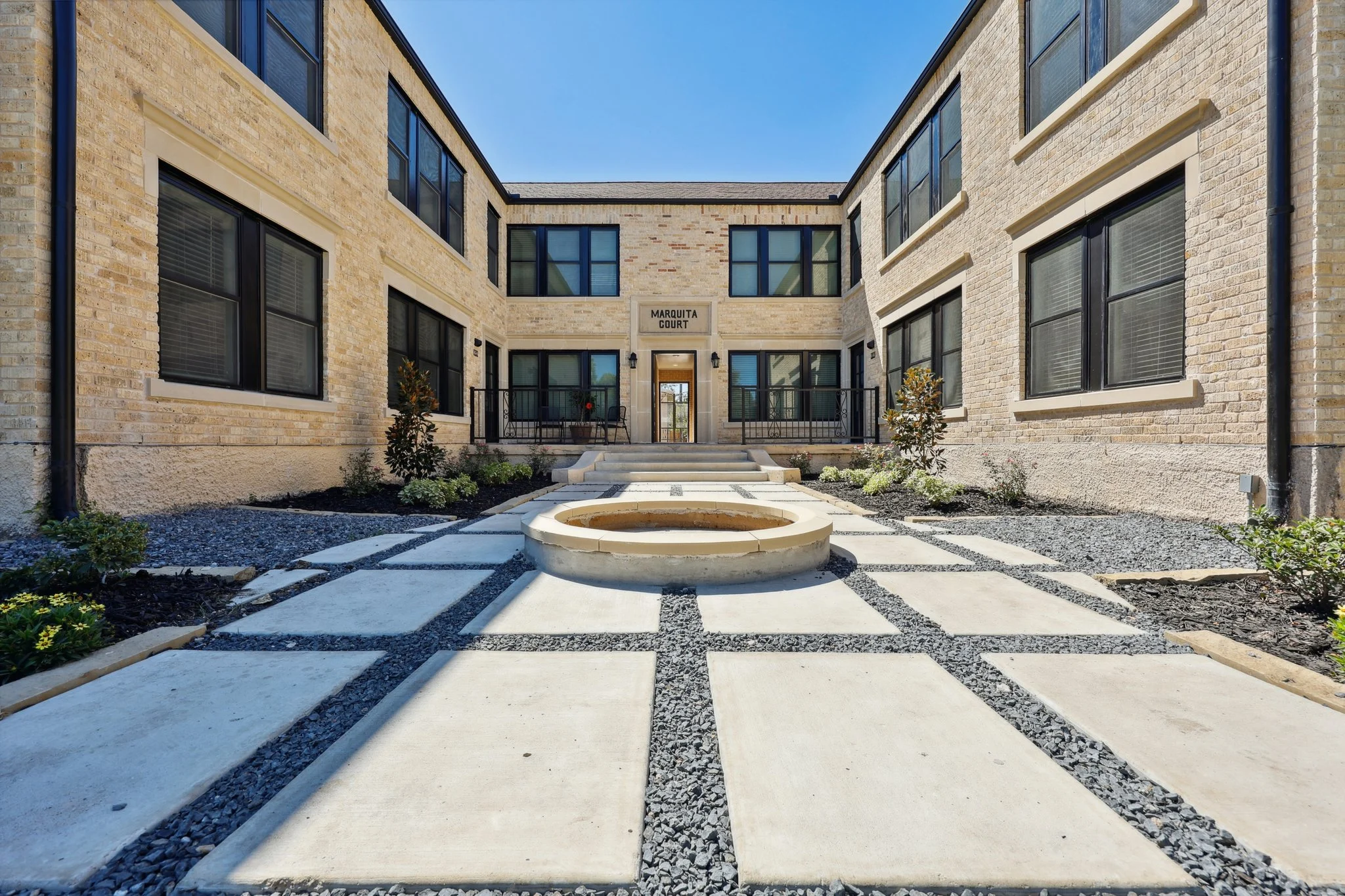 Modern Apartment Courtyard A modern apartment courtyard with stone buildings on either side, featuring a central circular fountain surrounded by a geometric pathway and gravel landscaping.