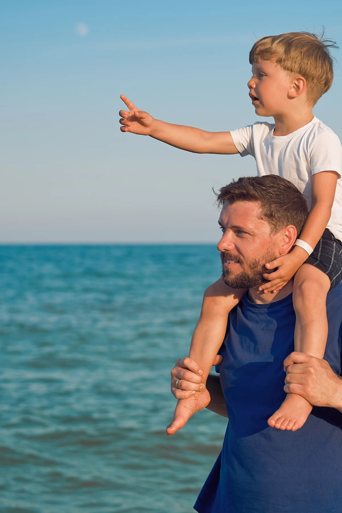 Father and Son Beach Day A father carrying his son on his shoulders at the beach, with the son pointing towards something in the distance.