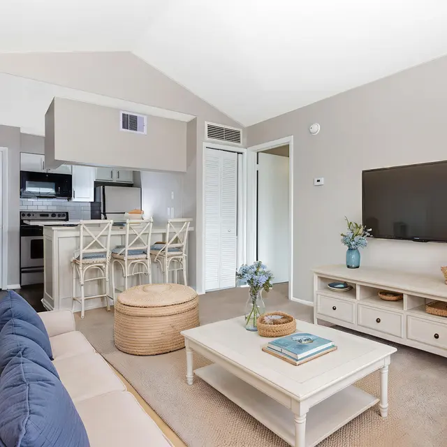 A modern living room with a beige sofa, a light wooden coffee table, a television mounted on the wall, and an open kitchen area in the background with bar stools.