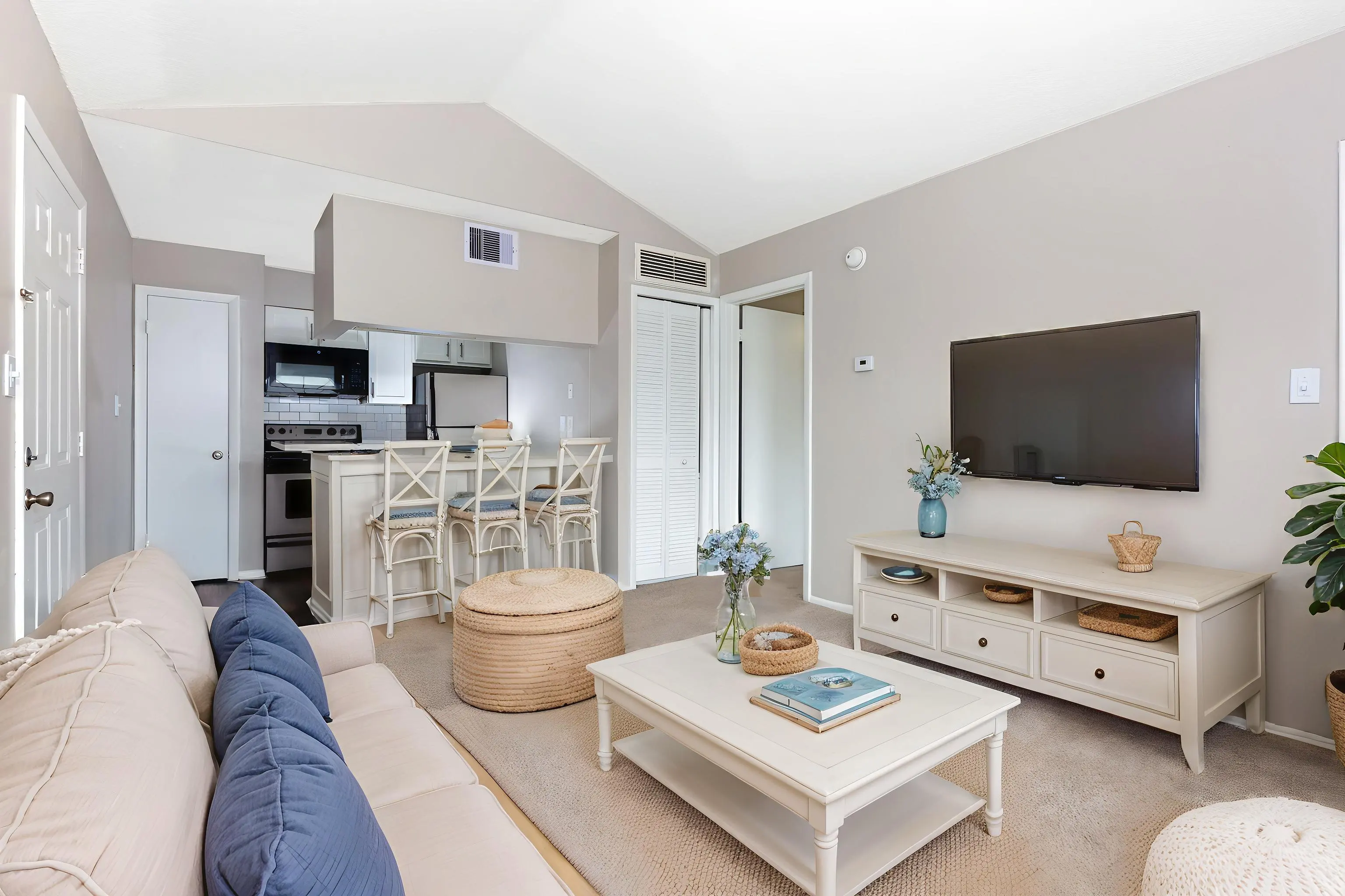 A modern living room with a beige sofa, a light wooden coffee table, a television mounted on the wall, and an open kitchen area in the background with bar stools.