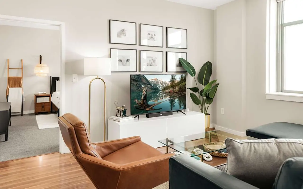 A stylish modern living room featuring a brown leather armchair, a white media console with a television, framed artwork on the wall, a green plant, and a cozy atmosphere.