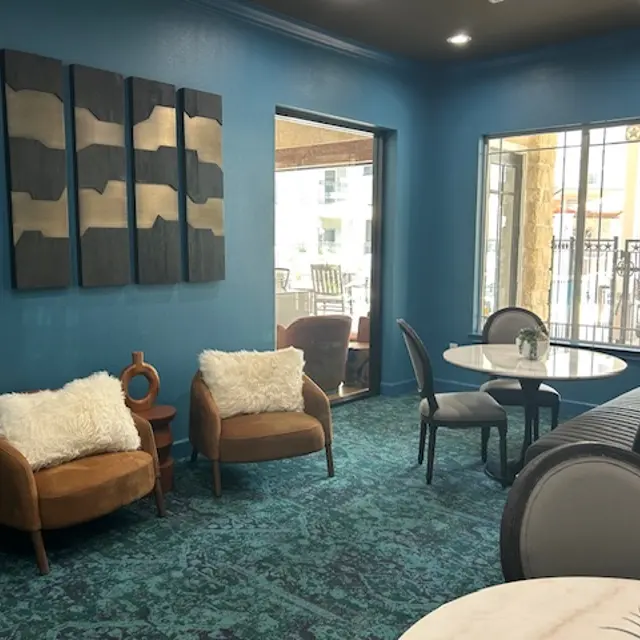 A modern lounge area featuring two stylish brown chairs with white fur throws, a small coffee table, and decorative wall art. The walls are painted a bright blue, and there is a view of a patio through large windows.