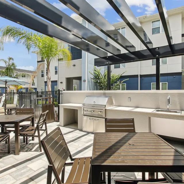 Outdoor patio area with a BBQ grill, tables, and chairs, surrounded by modern apartment buildings and palm trees.
