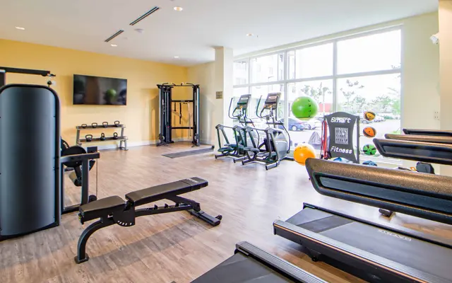 Interior view of a gym featuring various fitness equipment including treadmills, stationary bikes, and free weights. Large windows provide natural light.