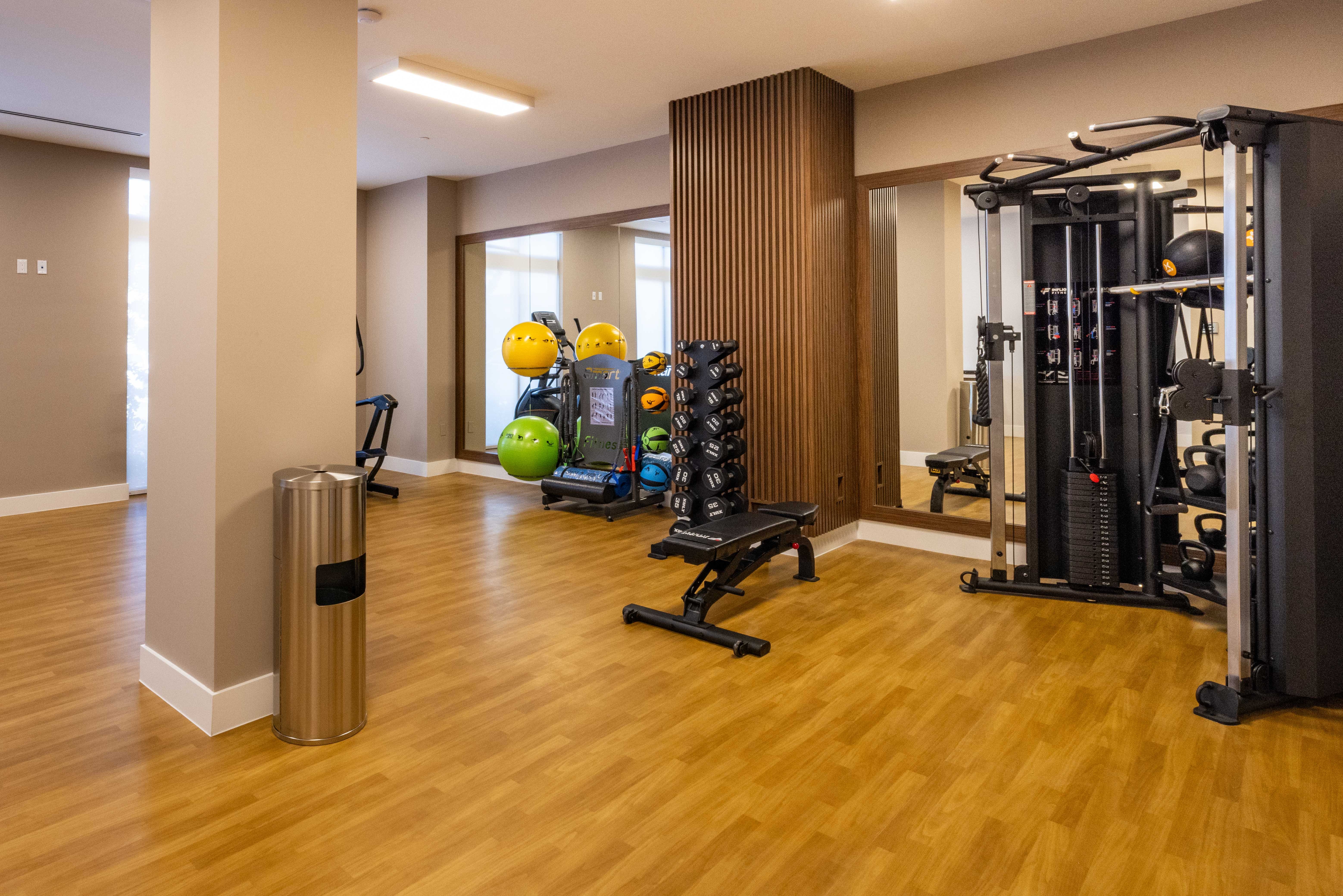 A modern gym interior featuring various fitness equipment, including weights and a multi-exercise machine. The flooring is wood-like, and the space has large windows for natural light.