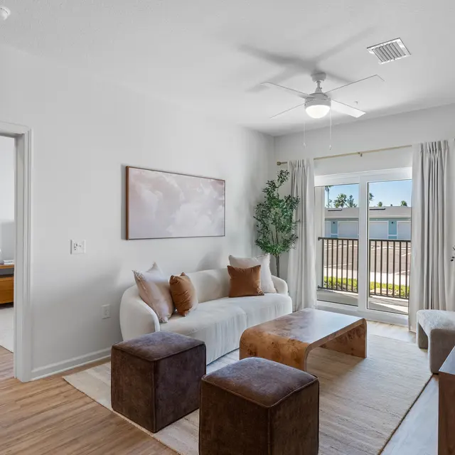 A modern living room featuring a white sofa with brown cushions, a wooden coffee table, and two brown ottomans. Large windows with curtains provide natural light, and decorative plants are placed in the corners. There are light wood floors, and a TV on a stand is visible.