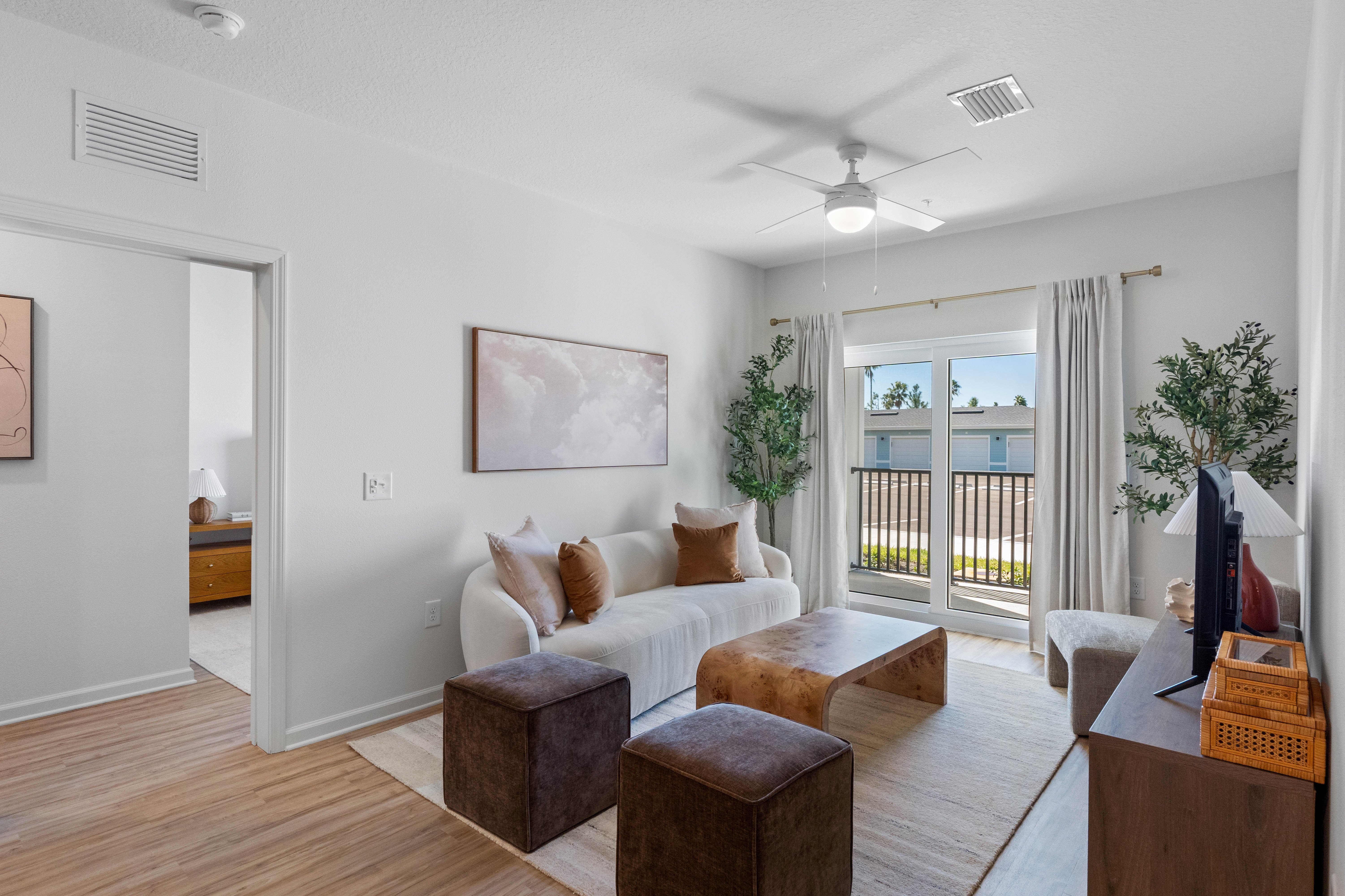 A modern living room featuring a white sofa with brown cushions, a wooden coffee table, and two brown ottomans. Large windows with curtains provide natural light, and decorative plants are placed in the corners. There are light wood floors, and a TV on a stand is visible.