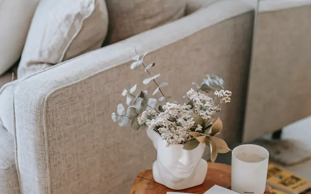A cozy living room corner featuring a gray sofa with cushions, a wooden side table holding a white vase with dried flowers, a candle, and a stack of magazines.