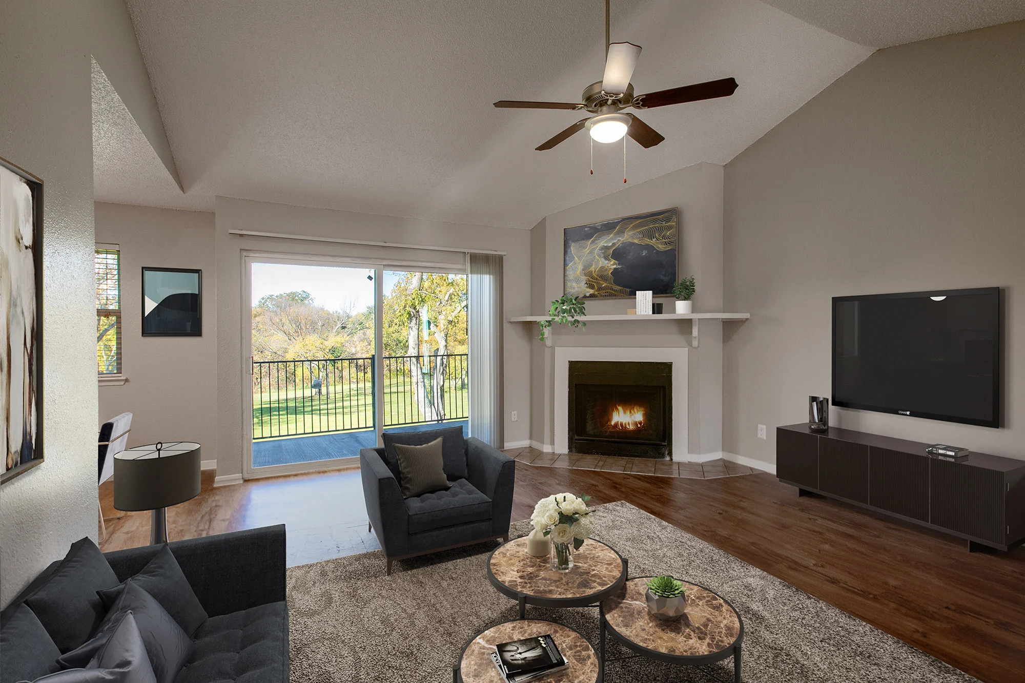A modern living room featuring a seating area with blue sofas and round coffee tables. A fireplace with a mantel is central, next to a wall-mounted television. Large windows overlook a green landscape.